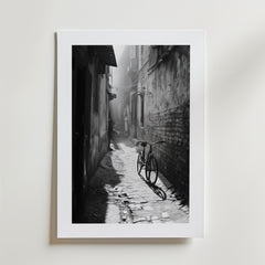 Quiet Echo Poster by Bozetto har ett svartvitt foto av en smal kullerstensgränd där solljuset kastar en cykels skugga genom dimman, tryckt på FSC-certifierat premiumpapper för en högkvalitativ finish.