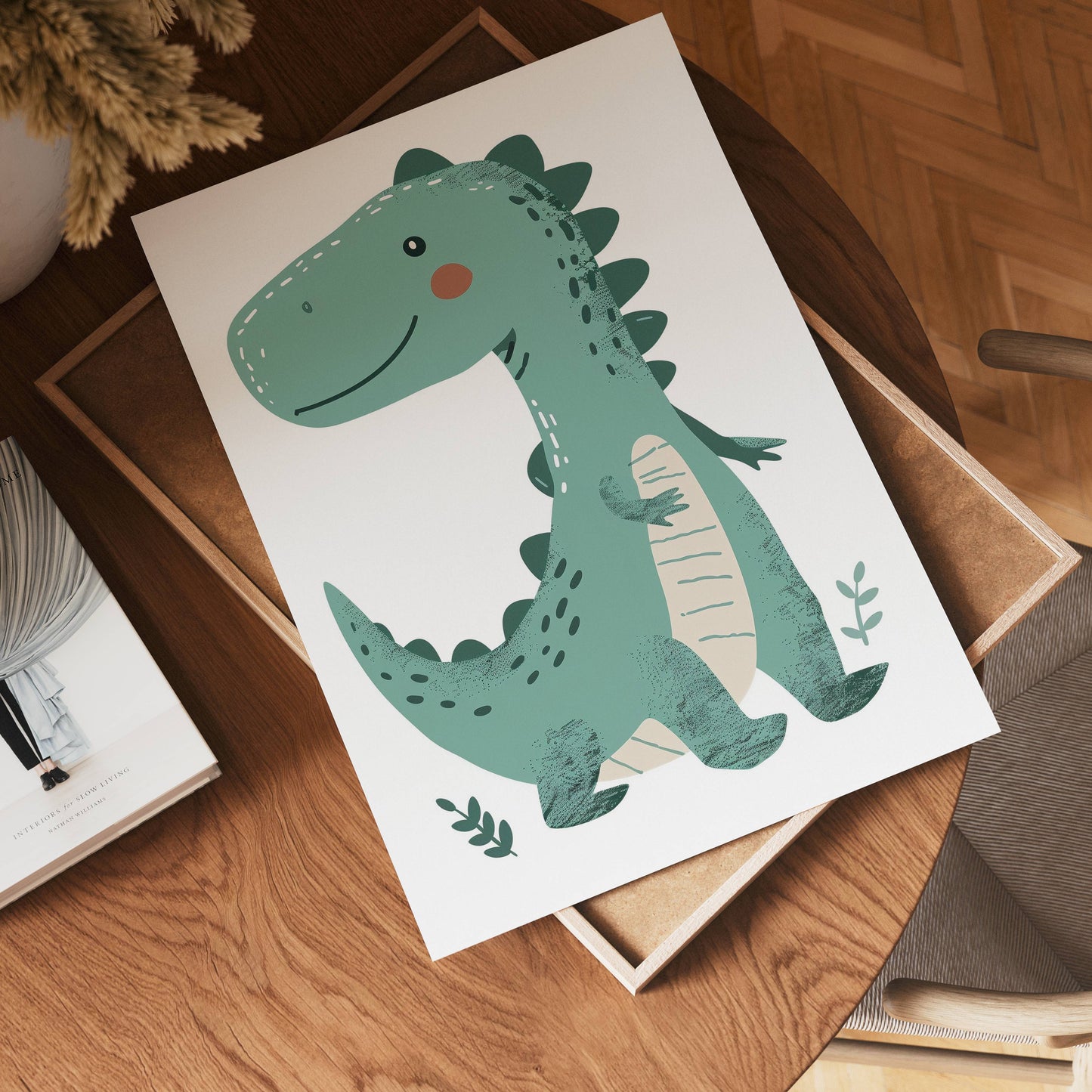 A cheerful, green cartoon dinosaur illustration from no-9 is displayed on white paper, resting on a wooden table beside a book, a chair, and a plant partly visible in the corner.